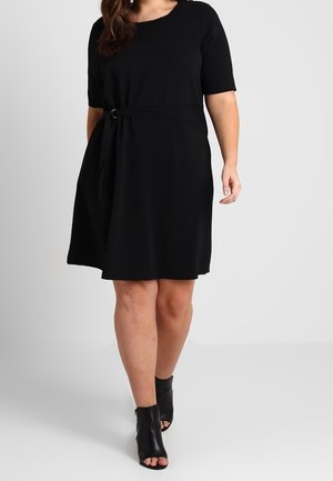 Femme portant une robe noire à manches courtes avec une ceinture nouée sur le côté et des bottines noires à bout ouvert, debout devant un fond gris uni.