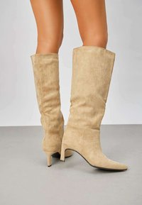 Bottes beige, montantes jusqu'au genou, en suede doux, avec un talon aiguille fin et un design à bout pointu. Texture lisse avec un minimum de détails.