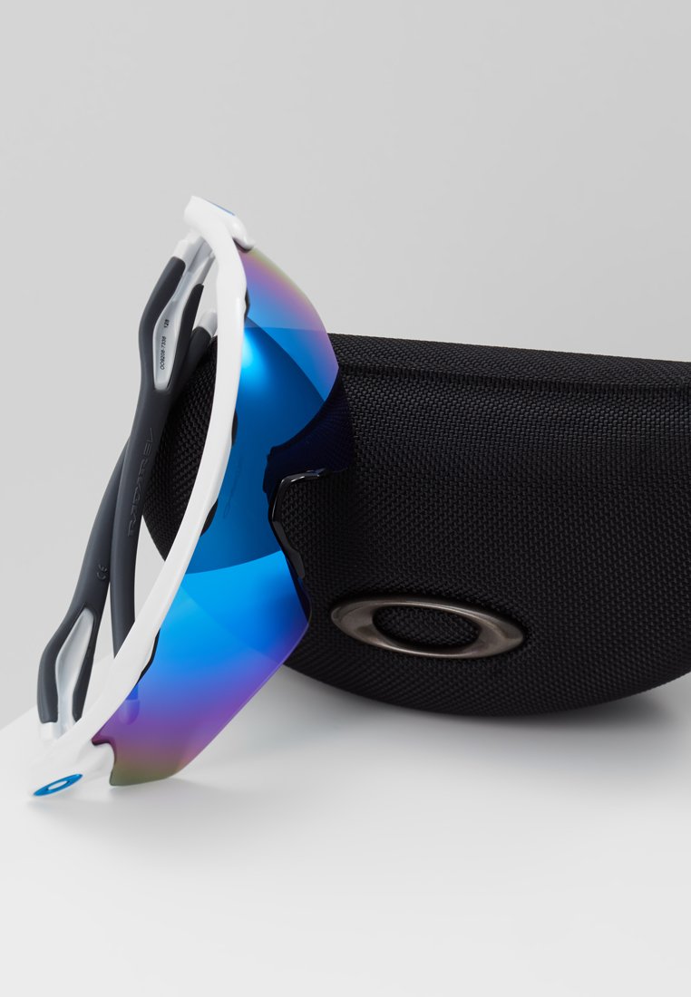 Occhiali da sole sportivi con grandi lenti riflettenti blu, montatura bianca e nera, e una custodia nera текстurizzata con logo.