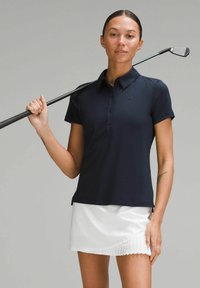 Mornarsko plavi polo majica s ovratnikom i tri gumba, uparen s bijelom plisiranom suknjom. Drži golf štap uz rame, glatka tekstura tkanine.