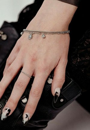 Hand with black and white marble-effect nails, moon design on ring finger, silver chain bracelet with charms, and thin ring, resting on black leather.