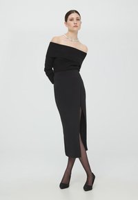 Top nero off-shoulder con texture a costina, abbinato a una gonna nera aderente con spacco alto fino alla coscia, e tacchi neri brillanti.