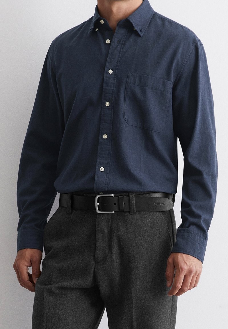 Homme portant une chemise bleu foncé boutonnée rentrée dans un pantalon gris avec une ceinture en cuir noire à boucle argentée.