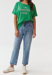 Camiseta verde de manga corta con el texto "MI CITA PARA SIEMPRE" en color rosa, combinada con unos vaqueros de denim azul claro de cintura alta y zapatillas beige.