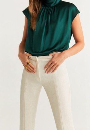 Kobieta w ciemnozielonej bluzce bez rękawów, wsuniętej w dopasowane, wysokiej talii spodnie w kolorze ecru, z rękami opartymi w pobliżu paska.