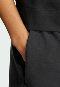 Čierna tkanina s hladkým povrchom. Detailný záber ruky v vrecku, ukazujúci prešitie na páse a uvoľnený strih materiálu.