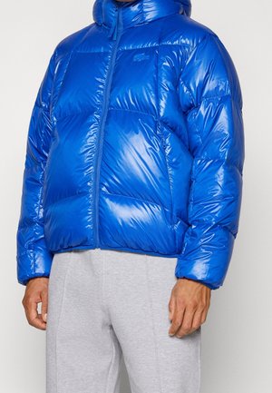 Homme portant une doudoune bleue brillante avec une fermeture éclair devant et un pantalon de survêtement gris, debout devant un fond uni.