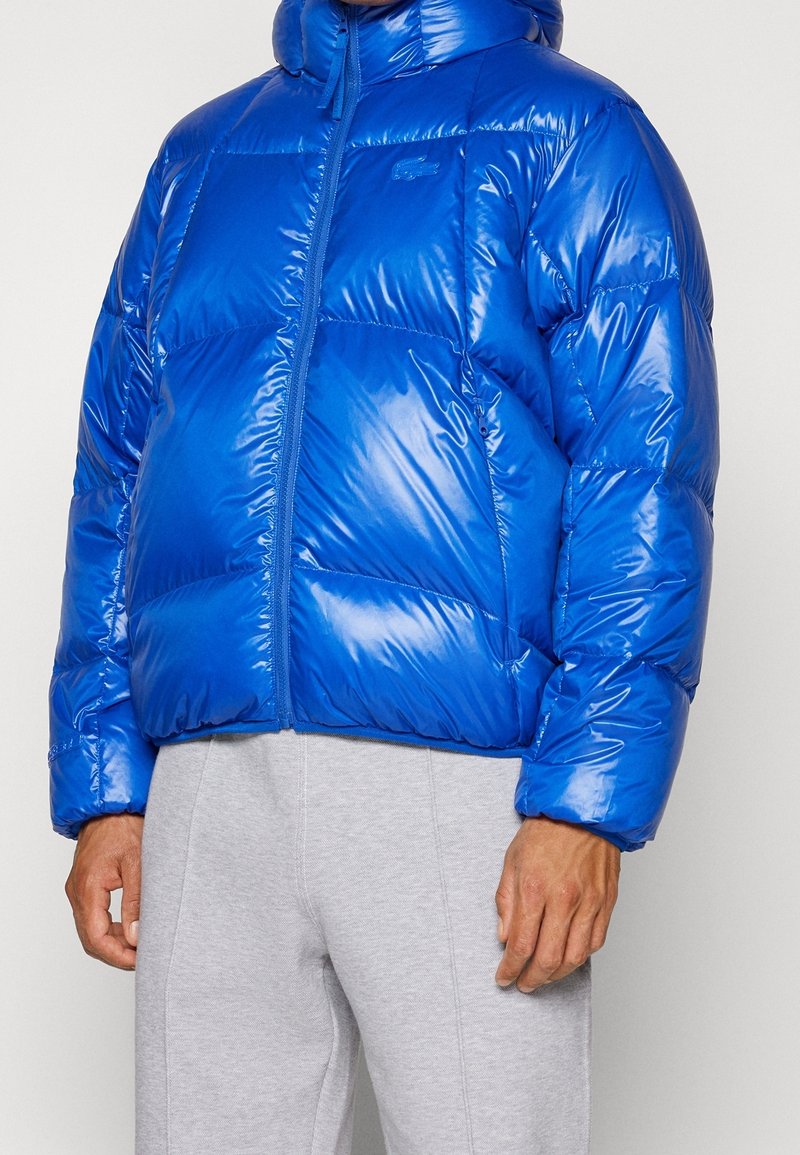 Homme portant une doudoune bleue brillante avec une fermeture éclair devant et un pantalon de survêtement gris, debout devant un fond uni.