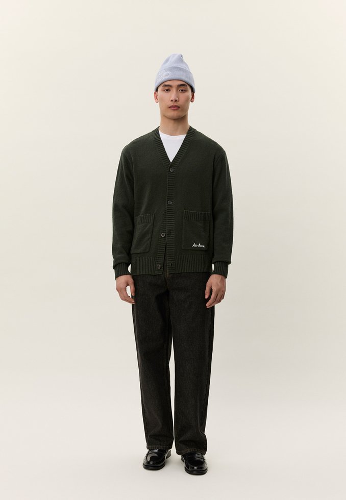 Άνδρας στέκεται φορώντας μια σκούρα πράσινη ζακέτα με κουμπιά, μαύρο παντελόνι, μαύρα παπούτσια, λευκή μπλούζα και ανοιχτό γκρι σκουφάκι, μπροστά σε απλό φόντο.