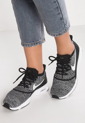 Feet wearing black and white knit Nike sneakers with laces, paired with cropped light blue denim jeans against a plain background.