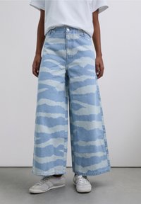 Light blue wide-leg jeans made of denim, featuring a wave pattern in a lighter shade. Paired with white sneakers, texture is matte.