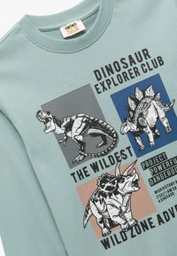 Miętowa bluza z długim rękawem z grafikami dinozaurów w czarnych, granatowych i różowych kwadratach, z napisem "Klub Odkrywców Dinozaurów" oraz "Najdzikszy".