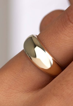Bague plaquée or avec un anneau lisse et arrondi. Présente une surface polie et une finition chaude et réfléchissante, mise en valeur sur le teint de la peau d'une main.