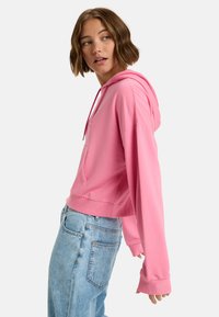 Krótka różowa bluza z kapturem, z możliwością regulacji, z kieszenią z przodu i długimi rękawami; gładka tekstura i swobodny krój, zestawiona z jasnoniebieskimi dżinsami.