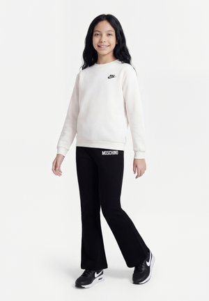 Fille debout, souriante, portant un sweat-shirt blanc Nike, un pantalon noir Moschino et des baskets noires Nike sur un fond blanc uni.