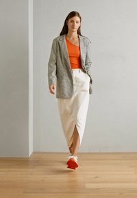 Blazer a quadri grigio e bianco, canotta arancione, gonna crema con spacco laterale e sneakers bianche con suole arancioni, posizionate su un pavimento in legno.
