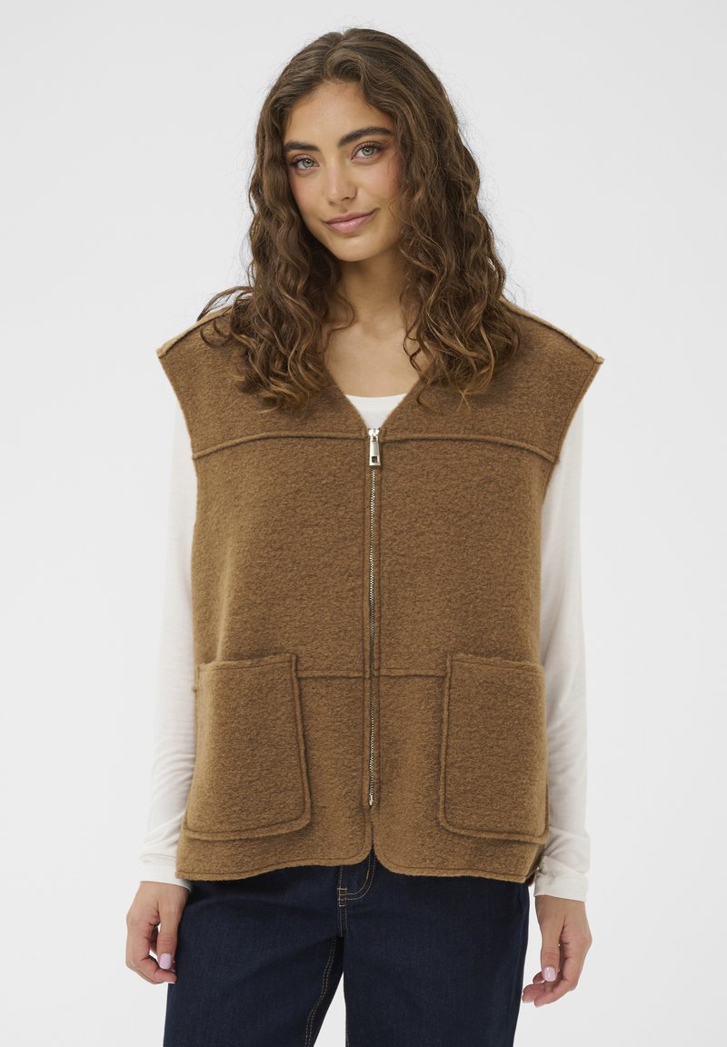 Mlada žena s kovrčavom kosom koja nosi smeđi prsluk bez rukava sa zipom preko bijele košulje dugih rukava i tamnoplavih traperica.