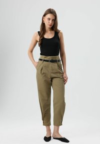 Zwarte aansluitende tanktop, olijfgroene broek met hoge taille en plooien, verstelbare zwarte riem en zwarte instappers, allemaal gepresenteerd op een neutrale achtergrond.