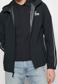 Černá větrovka s kapucí, s předním zipem, kontrastními bílými pruhy a elastickými manžetami. Nositá přes černé polo tričko.