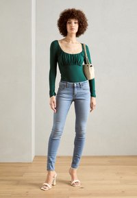 Grønn langermet bodysuit med rynket front, parret med lyseblå skinny jeans og nude blokkhelede sandaler. Liten beige håndveske.