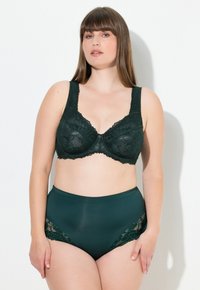 Tummanvihreä pitsinen rintaliivi leveillä nauhoilla ja kukkakuviolla, yhdistettynä korkeavyötäröisiin alushousuihin, joissa on pitsiyksityiskohtia sivuilla. Sujuva rakenne.