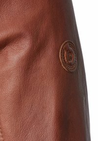 Cuero marrón con una superficie texturizada, que presenta un logotipo en relieve circular. El material parece suave y flexible. Los acentos son mínimos.