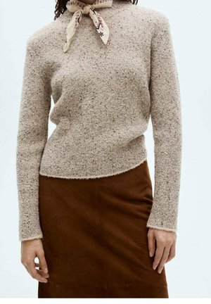 Kvinde iført en beige struktureret sweater, brun nederdel og et mønstret silketørklæde bundet rundt om halsen.