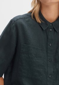 Camicia di lino verde scuro con maniche corte, una chiusura con bottoni sul davanti, colletto e due tasche sul petto. Tessuto morbido con una finitura leggermente stropicciata.