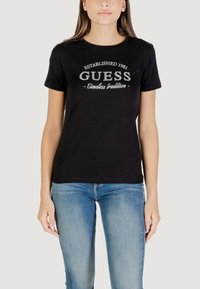T-shirt di cotone nero con logo "GUESS" in argento, scollatura rotonda, maniche corte e vestibilità casual, abbinato a jeans azzurri chiari.
