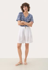 Blå kortärmad topp med vitt blommönster, tillsammans med vita linnebyxor med en knapp. Modell står med armarna i kors och har på sig flip-flops.