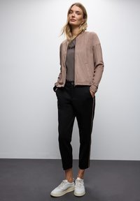 Tan mockaläder bomberjacka, grå t-shirt med svart tryck, svarta tapered byxor med bruna sidostreck och vita sneakers.