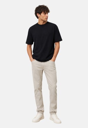 Hombre con camisa negra de manga corta acanalada, pantalones beige claros y zapatillas blancas, de pie con las manos en los bolsillos, mirando hacia un lado.