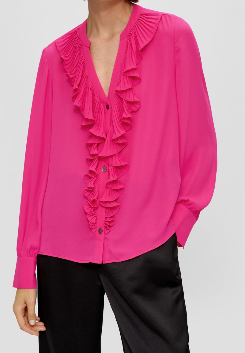 Modell klädd i en ljusrosa blus med volangdetaljer fram, kombinerad med svarta satinbyxor, poserande mot en neutral bakgrund.