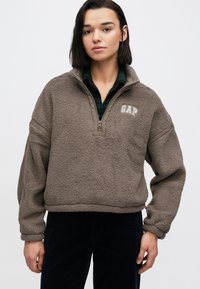 Brązowy polarowy pulower z wysokim kołnierzem, zamkiem na ćwierć długości i logo GAP. Charakteryzuje się szerokimi rękawami i luźnym krojem, zestawiony z ciemnymi spodniami.