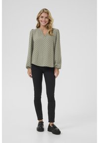 Licht olijfgroene blouse met zwarte stippen, v-hals, lange pofmouwen en zachte stof, gedragen met zwarte skinny jeans en zwarte schoenen.