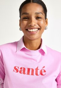 Lyserød t-shirt med rød "santé" print, lagdelt over en hvid og lyserød stribet skjorte med krave. Modellen bærer runde, glitrende øreringe.