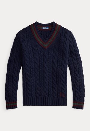 Granatowy sweter w warkocze z dekoltem w serek, szare spodnie, brązowe skórzane buty oraz kraciasty krawat. Subtelna tekstura i kontrastujące akcenty swetra.