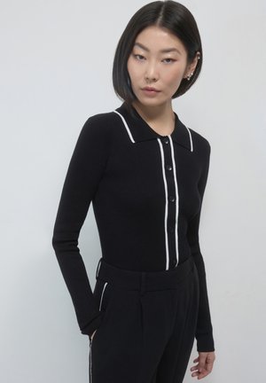 Schwarzes geripptes Langarm-Polohemd mit einem weißen Kragen und Knöpfen auf der Vorderseite. Das Design umfasst kontrastierende weiße Besätze entlang der Kanten.