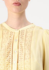 Blusa amarilla con paneles de encaje, botones de nácar y tejido texturizado. Tiene un escote redondo y mangas abullonadas.
