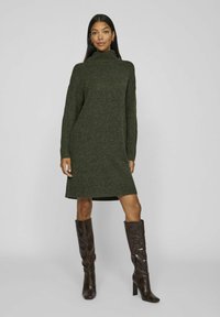 Vestito maglione verde scuro con colletto alto, maniche lunghe e orli a coste. Abbinato a stivali alti fino al ginocchio con motivo a pelle di serpente marrone.