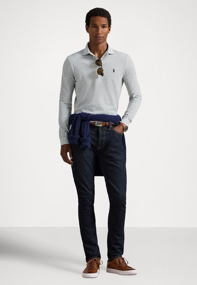 Ανοιχτό γκρι μακρυμάνικο πόλο πουκάμισο, σκούρο τζιν, καφέ αθλητικά παπούτσια, μπλε πουλόβερ δεμένο γύρω από τη μέση, γυαλιά ηλίου και ρολόι χειρός.