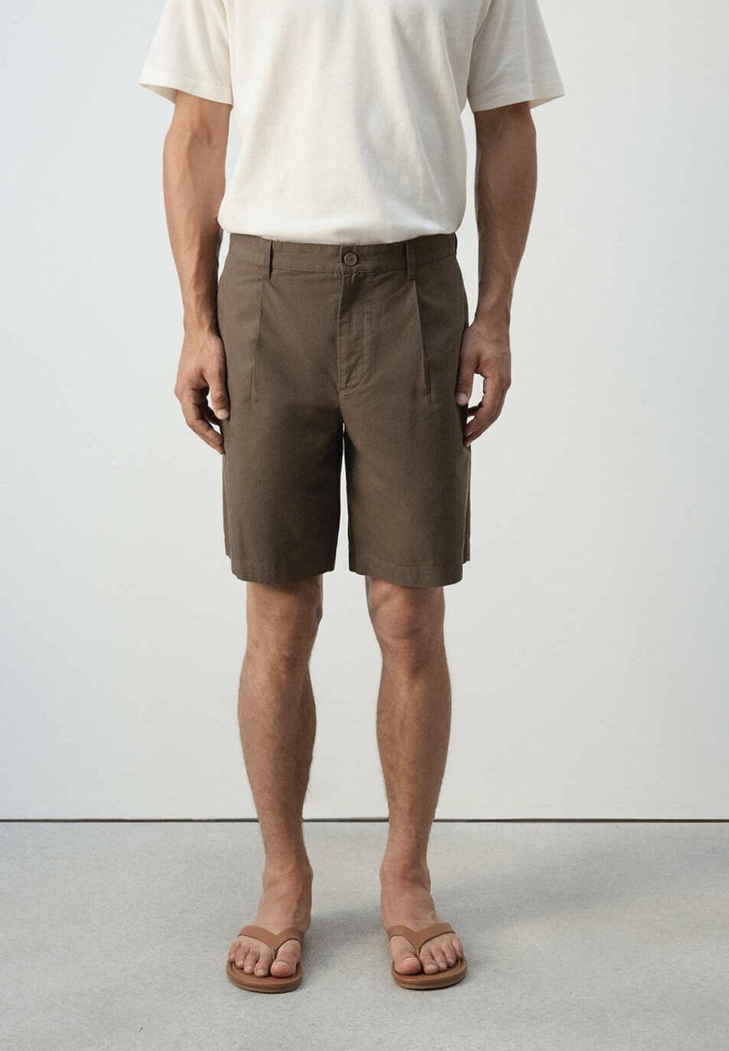 Homme portant un short brun jusqu'aux genoux, une chemise blanche à manches courtes et des sandales brunes, debout devant un fond uni.