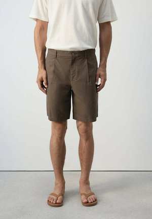 Homme portant un short brun jusqu'aux genoux, une chemise blanche à manches courtes et des sandales brunes, debout devant un fond uni.