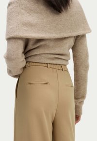 Beige pletený svetr s off-shoulder designem spárovaný s vysokými bežovými kalhotami, které mají dvě zadní kapsy a hladkou texturu.