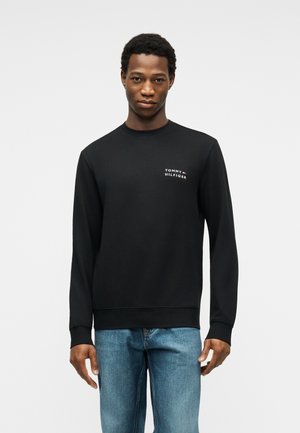 Man wearing a black Tommy Hilfiger sweatshirt and blue jeans, standing against a plain light gray background.