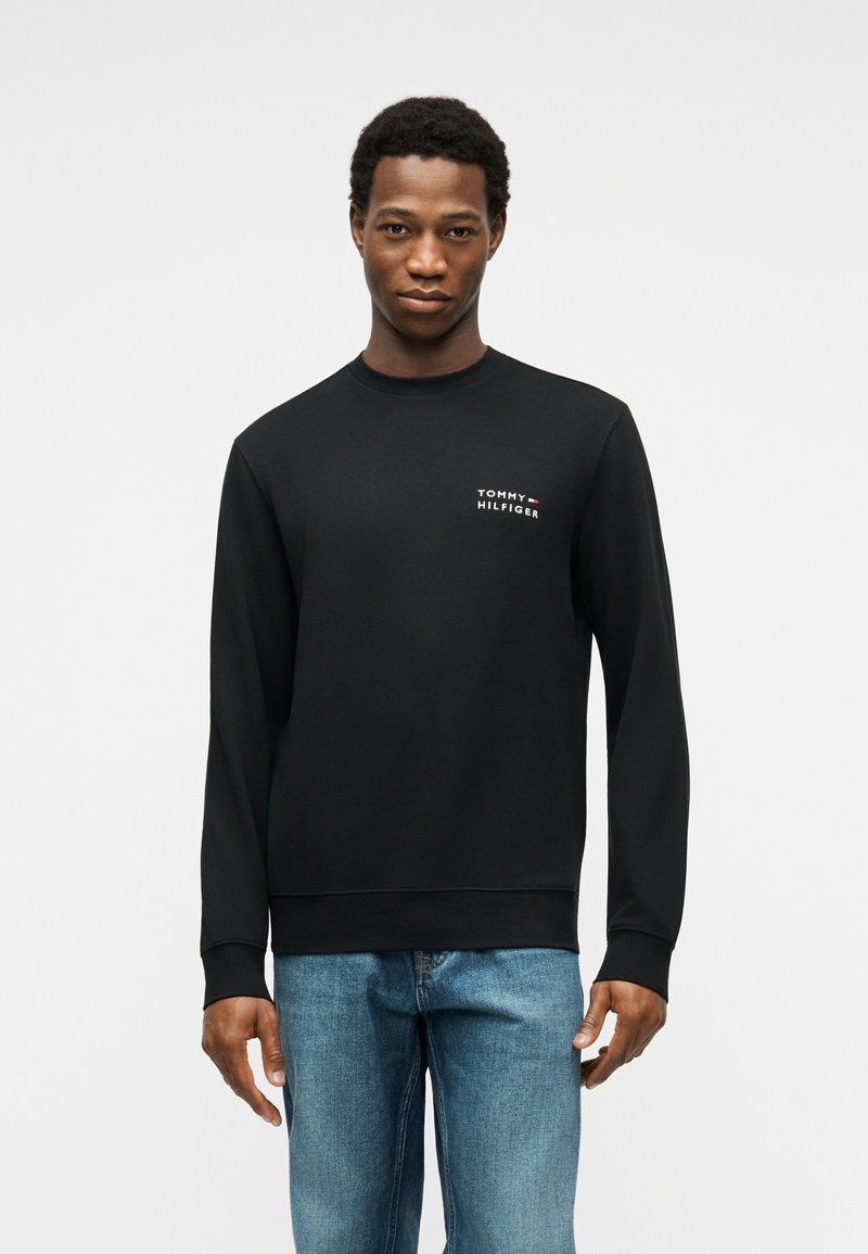 Άνδρας με μαύρο φούτερ Tommy Hilfiger και μπλε τζιν, στέκεται μπροστά σε απλό ανοιχτό γκρι φόντο.