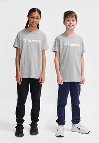 Tricouri gri din bumbac cu logo alb "hummel", asociate cu pantaloni de trening negri și bleumarin. Amândoi poartă încălțăminte sportivă. Design simplu și casual.