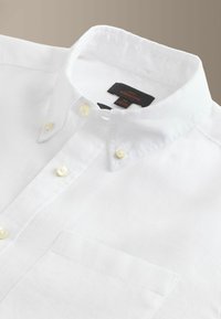 Chemise blanche à col, poche avant et boutons couleur crème, posée à plat sur un fond neutre.