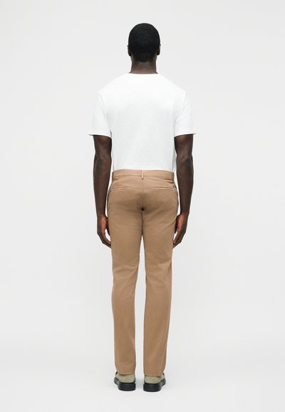 Homme debout, de dos, portant une chemise blanche à manches courtes, un pantalon beige et des chaussures de couleur claire sur un fond uni.