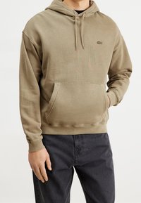Felpa con cappuccio di color oliva terroso, realizzata in tessuto morbido, presenta una tasca a marsupio frontale e un logo ricamato sul petto.